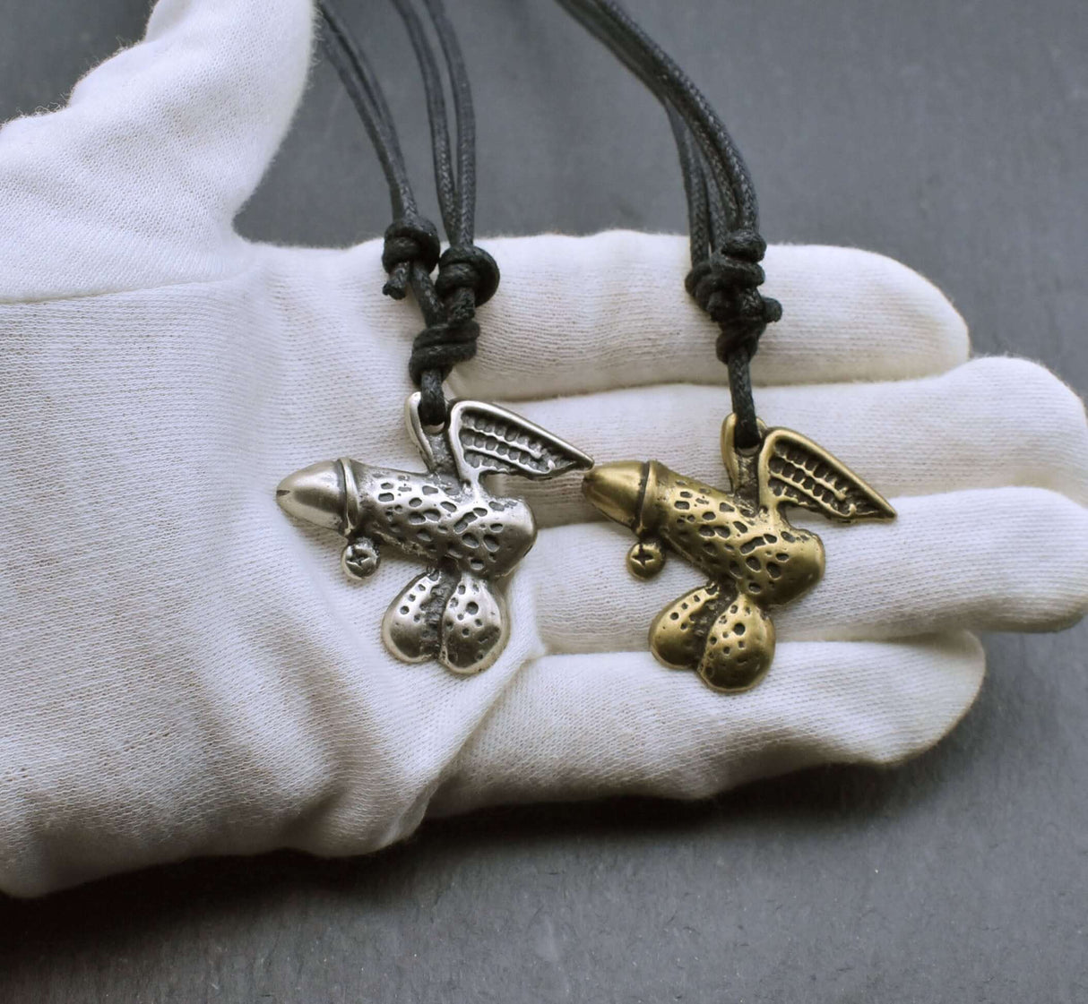 Geflügeltes Phallus-Amulett als humorvolle "Pimmelkette" aus ZAMAK in Messing- und Silberfarben auf der Hand