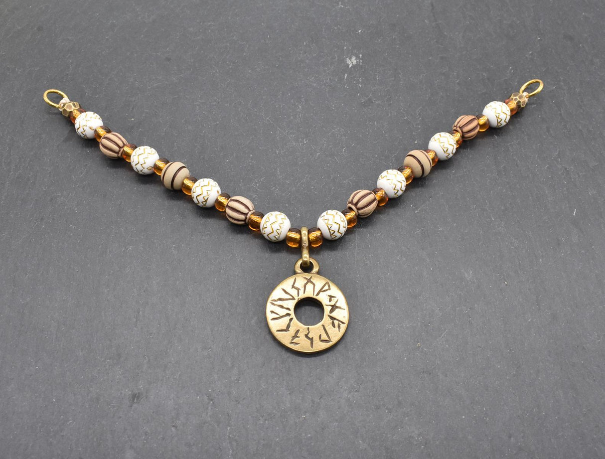 Handgefertigte Runen Fibelkette von BELANAS SCHATZKISTE mit hellbraunen und weißen Acrylperlen, goldfarbenen Mustern, braunen Goldfolienperlen und einem runden Runenamulett aus ZAMAK. Elegantes Design mit mystischem Wikinger-Charme.
