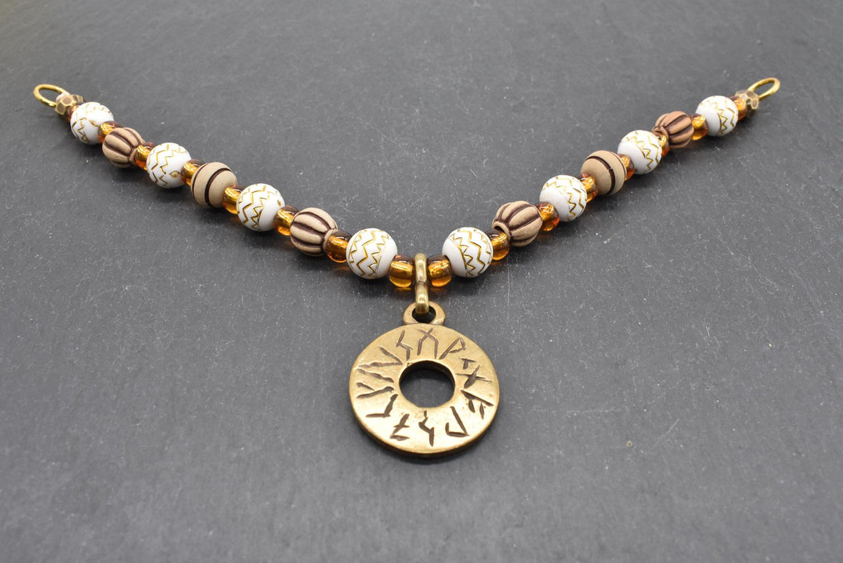 Handgefertigte Runen Fibelkette von BELANAS SCHATZKISTE mit hellbraunen und weißen Acrylperlen, goldfarbenen Mustern, braunen Goldfolienperlen und einem runden Runenamulett aus ZAMAK. Elegantes Design mit mystischem Wikinger-Charme.