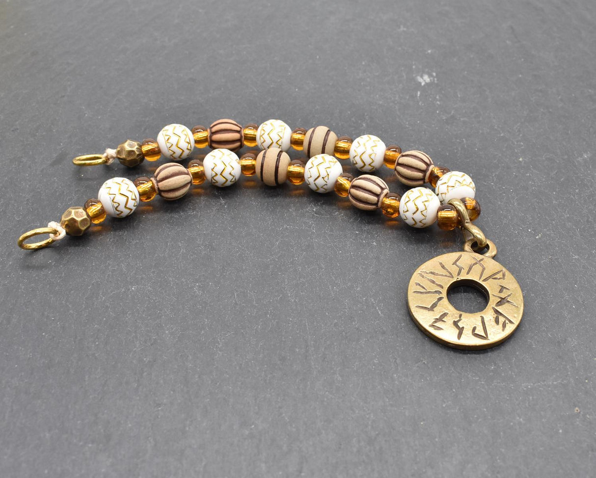 Handgefertigte Runen Fibelkette von BELANAS SCHATZKISTE mit hellbraunen und weißen Acrylperlen, goldfarbenen Mustern, braunen Goldfolienperlen und einem runden Runenamulett aus ZAMAK. Elegantes Design mit mystischem Wikinger-Charme.