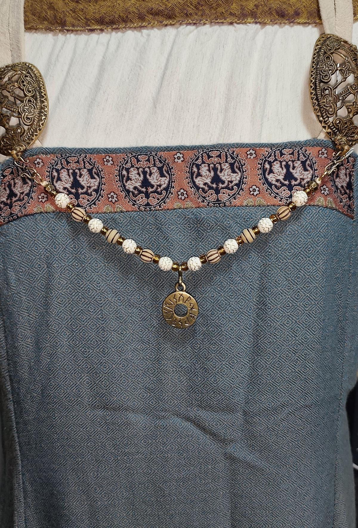 Tragebild am Wikingerkleid der Handgefertigte Runen Fibelkette von BELANAS SCHATZKISTE mit hellbraunen und weißen Acrylperlen, goldfarbenen Mustern, braunen Goldfolienperlen und einem runden Runenamulett aus ZAMAK. Elegantes Design mit mystischem Wikinger-Charme.