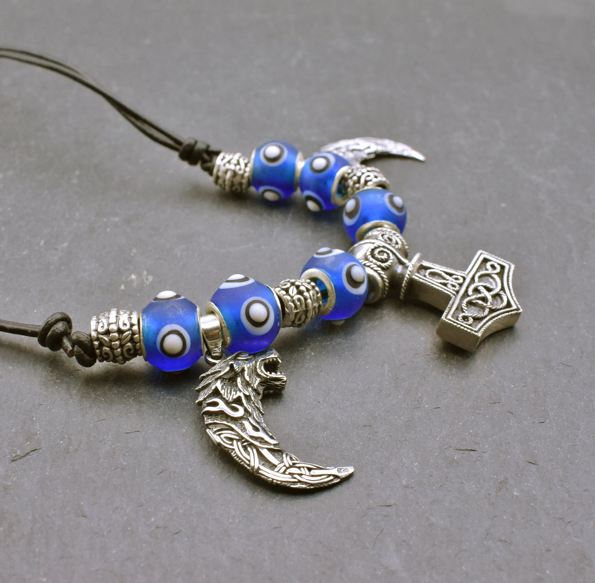 Mjölnir-Kette „Ragnar“ kombiniert drei Anhänger aus 925er Sterling Silber mit kräftig blauen, handgefertigten Glasperlen und schwarzem Lederband.
Im Mittelpunkt sitzt ein reich verzierter Thorshammer. Zwei gebogene, zahnförmige Anhänger mit plastisch ausgearbeiteten Wolfsköpfen flankieren den Mjölnir.