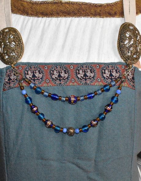 Tragebild der Handgefertigte Fibelkette aus blauen und goldbraunen Glasperlen mit bronzefarbenen Metallringen, perfekt für Wikingerkleidung und Reenactment, gefertigt von Belanas Schatzkiste.