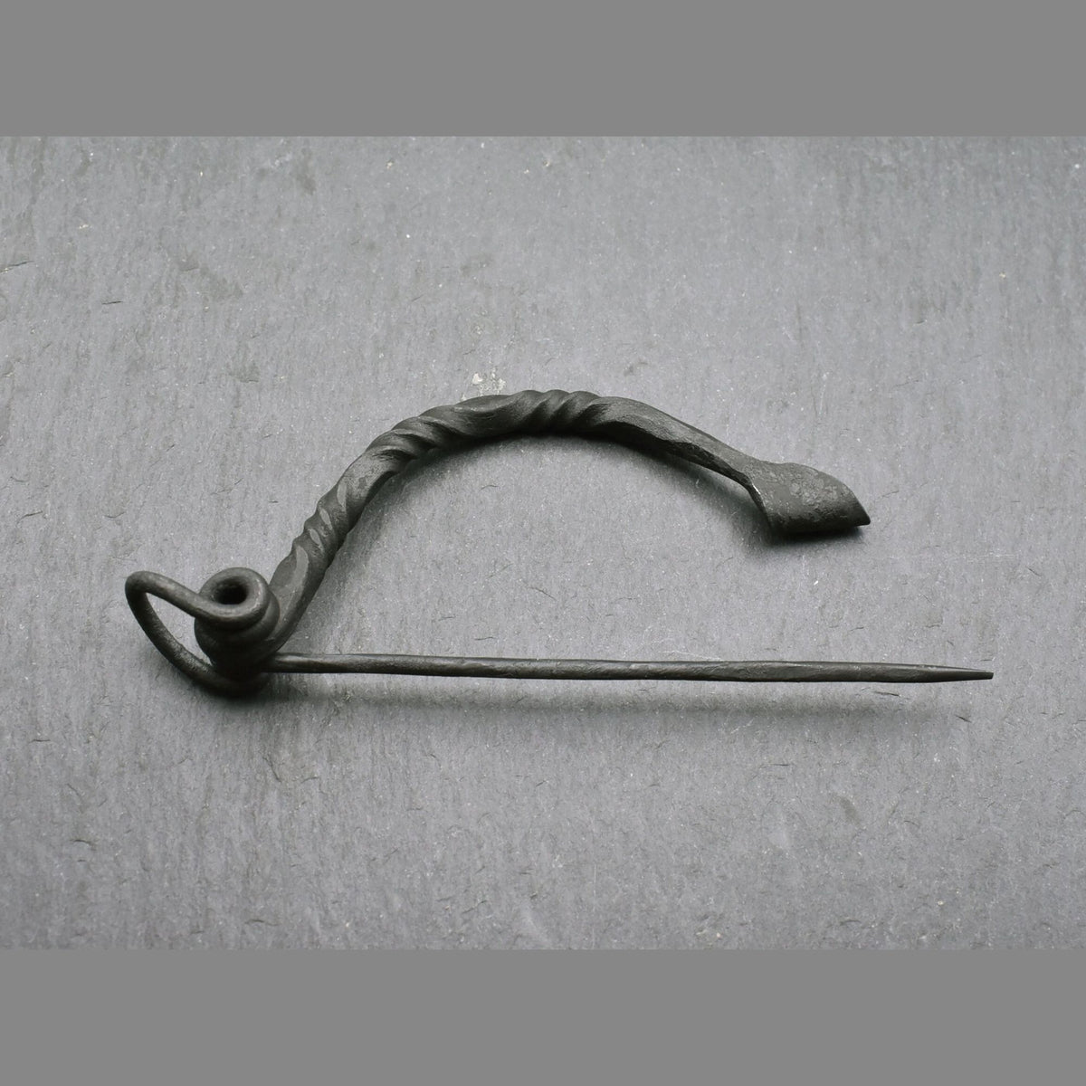 Keltische Bügelfibel aus Eisen – handgeschmiedete Latène-Fibel

Diese keltische Bügelfibel aus Eisen ist von Hand geschmiedet und orientiert sich an historischen Fibelformen der Latènezeit. Der kräftige Bügel ist teilweise tordiert und geht in eine Spiralkonstruktion mit langer Nadel über. Am anderen Ende befindet sich der Nadelhalter, in den die Spitze zum Verschließen des Stoffes eingelegt wird.