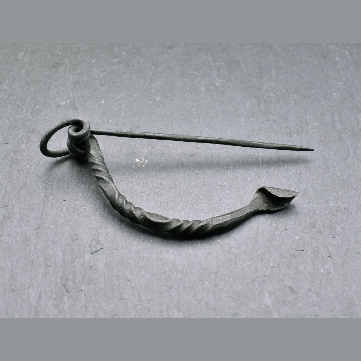 Keltische Bügelfibel aus Eisen – handgeschmiedete Latène-Fibel

Diese keltische Bügelfibel aus Eisen ist von Hand geschmiedet und orientiert sich an historischen Fibelformen der Latènezeit. Der kräftige Bügel ist teilweise tordiert und geht in eine Spiralkonstruktion mit langer Nadel über. Am anderen Ende befindet sich der Nadelhalter, in den die Spitze zum Verschließen des Stoffes eingelegt wird.