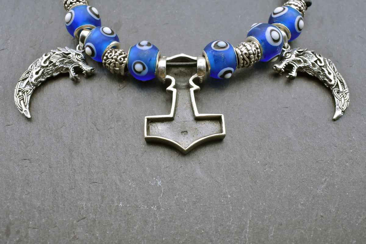 Rückseite mit 925er Stempel des Mjölnir Anhängers der Mjölnir-Kette „Ragnar“ kombiniert drei Anhänger aus 925er Sterling Silber mit kräftig blauen, handgefertigten Glasperlen und schwarzem Lederband.

Im Mittelpunkt sitzt ein reich verzierter Thorshammer. Zwei gebogene, zahnförmige Anhänger mit plastisch ausgearbeiteten Wolfsköpfen flankieren den Mjölnir.
