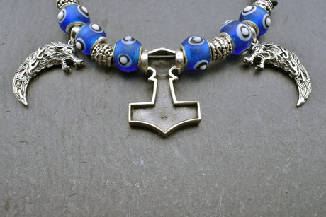 Rückseite mit 925er Stempel des Mjölnir Anhängers der Mjölnir-Kette „Ragnar“ kombiniert drei Anhänger aus 925er Sterling Silber mit kräftig blauen, handgefertigten Glasperlen und schwarzem Lederband.

Im Mittelpunkt sitzt ein reich verzierter Thorshammer. Zwei gebogene, zahnförmige Anhänger mit plastisch ausgearbeiteten Wolfsköpfen flankieren den Mjölnir.