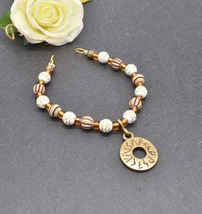 Handgefertigte Runen Fibelkette von BELANAS SCHATZKISTE mit hellbraunen und weißen Acrylperlen, goldfarbenen Mustern, braunen Goldfolienperlen und einem runden Runenamulett aus ZAMAK. Elegantes Design mit mystischem Wikinger-Charme.