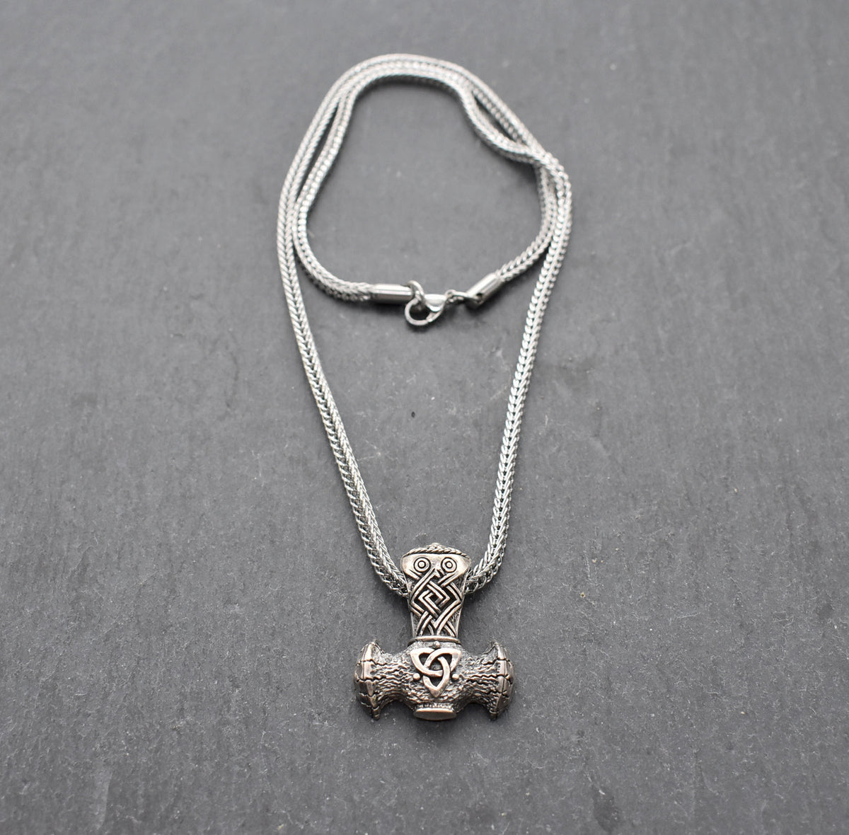 Thorshammer-Kette aus 925er Sterling Silber

Der massiv gearbeitete Thorshammer aus 925er Sterling Silber ist der Mittelpunkt dieser nordisch inspirierten Halskette.

Verschlungene Flechtornamente verzieren den Schaft des Mjölnir, während auf dem Hammerkopf eine Triskele sitzt. Die Vertiefungen sind dunkel abgesetzt und lassen die plastischen Details des Silberanhängers deutlich hervortreten.

Kombiniert ist der etwa 8,5 g schwere Anhänger mit einer 56 cm langen Fuchsschwanzkette aus Edelstahl. Geschlossen 
