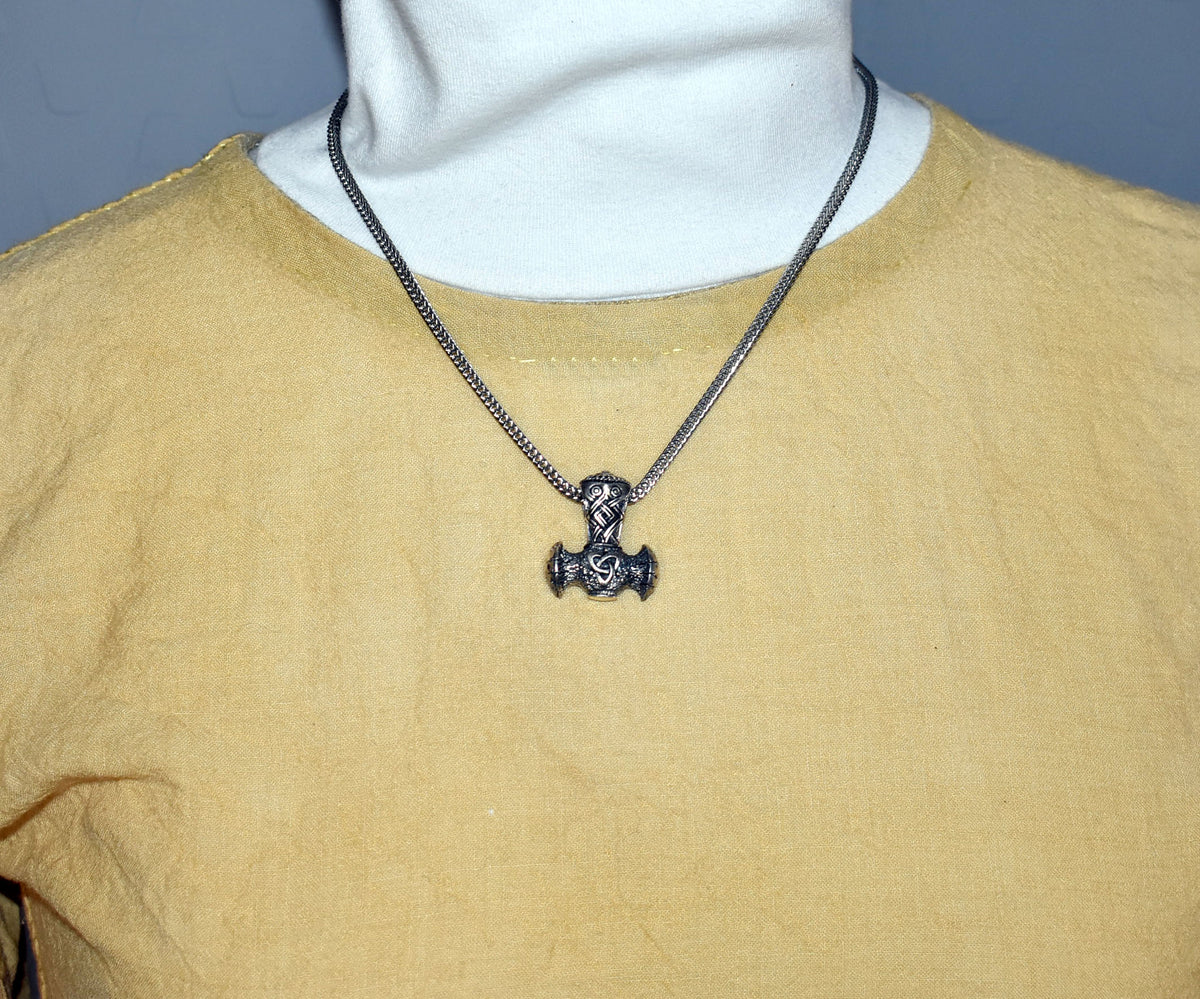 Tragebild der Thorshammer-Kette aus 925er Sterling Silber

Der massiv gearbeitete Thorshammer aus 925er Sterling Silber ist der Mittelpunkt dieser nordisch inspirierten Halskette.

Verschlungene Flechtornamente verzieren den Schaft des Mjölnir, während auf dem Hammerkopf eine Triskele sitzt. Die Vertiefungen sind dunkel abgesetzt und lassen die plastischen Details des Silberanhängers deutlich hervortreten.

Kombiniert ist der etwa 8,5 g schwere Anhänger mit einer 56 cm langen Fuchsschwanzkette aus Edelstahl