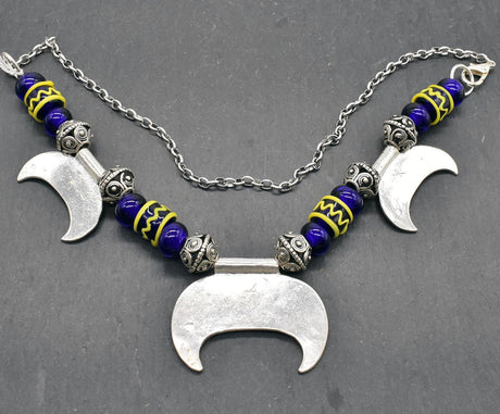 Rückseite der Lunula Fibelkette INGRID mit dunkelblauen Glasperlen mit gelbem Muster, sowie silbernen Perlen und Anhängern auf einer Schieferplatte 